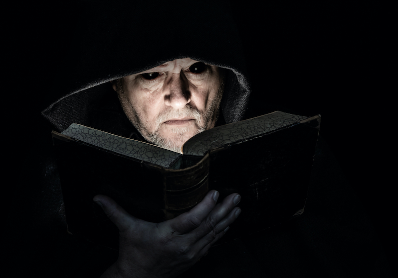 A scary looking man in a black robe reads a book in a dimly lit room