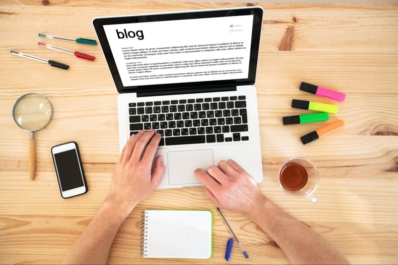 overhead view of a desk with laptop, phone, pens, and paper. The laptop screen says "blog". 