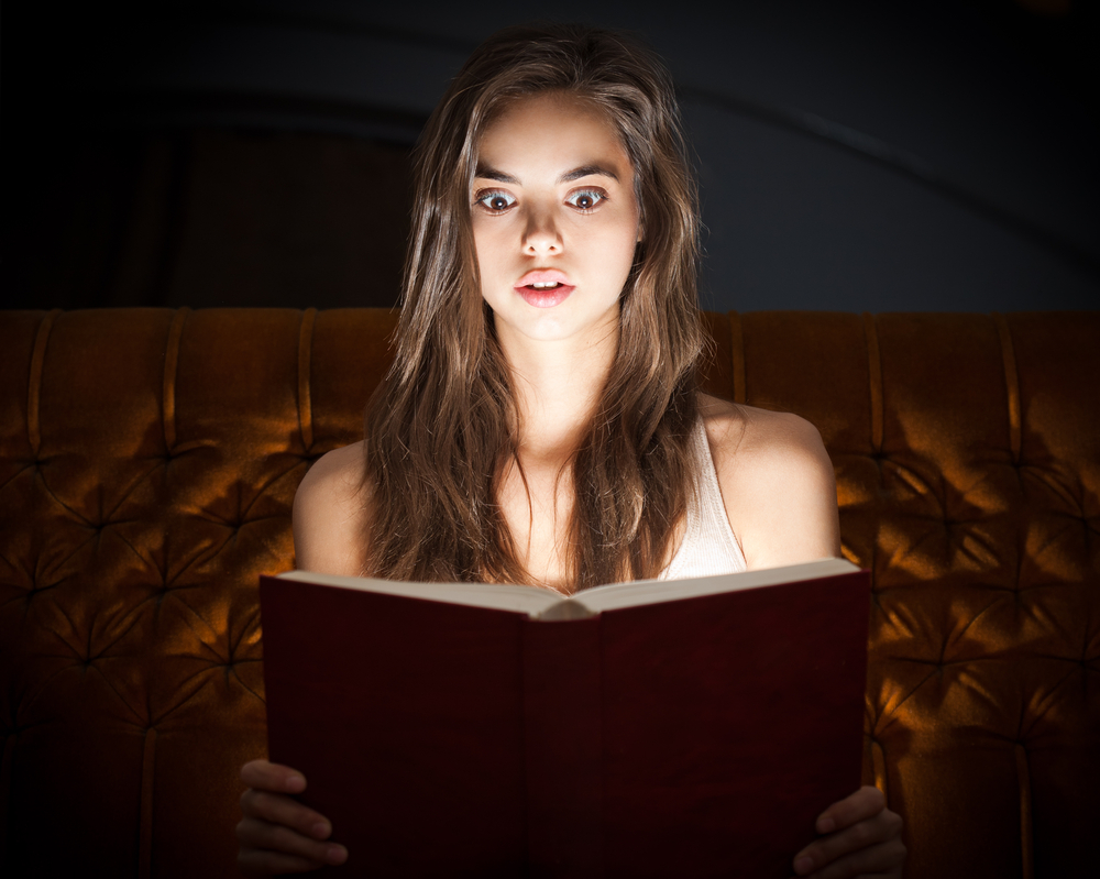 Young brunette woman looking shocked while reading a book